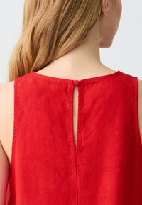 Rotes ärmelloses Top mit einem Rückenschlitz, Knopfverschluss und einem weichen, strukturierten Leinenstoff. Klare Linien und einfaches Design.