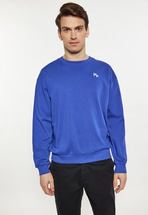 Ung mand står iført en blå sweatshirt med teksten "Mr." på brystet og sorte bukser, foran en ensfarvet lys baggrund.