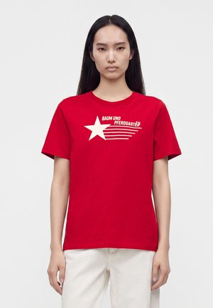 Femme aux longs cheveux noirs porte un t-shirt rouge avec une étoile blanche et le texte "Baum und Pferdgarten" ainsi qu'un pantalon blanc sur un fond uni.