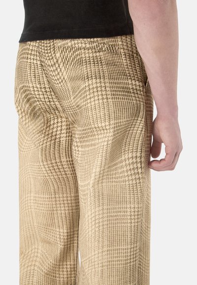 Une personne porte un pantalon à motif pied-de-poule beige et marron avec des poches arrière et un haut noir, debout devant un fond clair.