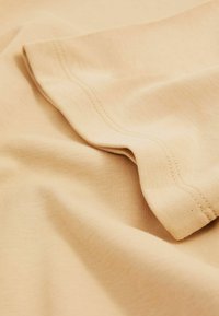 Beige puuvillakangas, jossa on sileä pinta, hienovaraiset poimukkeet ja päällekkäinen taitos. Ompeleet ovat näkyvissä.