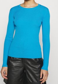 Femme portant un haut à manches longues côtelé bleu vif et un short en cuir noir, sur un fond clair uni.