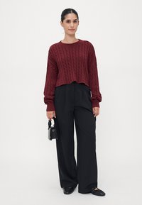 Burgundy cable-knit cropped sweater with long sleeves paired with black wide-leg trousers. Model holds a small black bag and wears flat shoes.