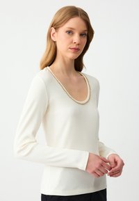 Kobieta z blond włosami, ubrana w biały, prążkowany top z długim rękawem z złotym łańcuszkiem wokół okrągłego dekoltu, stojąca na jednolitym tle.