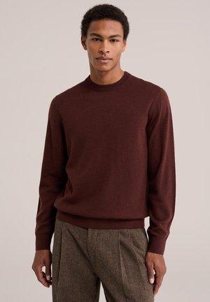 Jeune homme portant un pull bordeaux à manches longues et un pantalon marron à motifs, debout devant un fond clair uni.