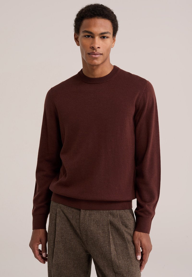 Jeune homme portant un pull bordeaux à manches longues et un pantalon marron à motifs, debout devant un fond clair uni.