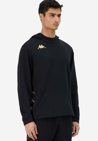 Felpa nera con cappuccio, tasca frontale, logo dorato sulla spalla sinistra e dettagli dorati lungo i lati. Tessuto morbido, vestibilità rilassata.