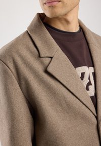 Cappotto marrone in misto lana con rever a punta, caratterizzato da una texture morbida. Indossato sopra una felpa marrone scuro con un motivo beige chiaro.