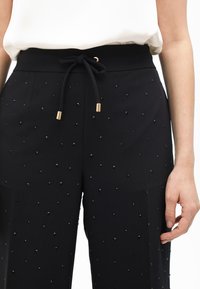 Čierne nohavice s elastickým pásom so šnúrkou, ktoré majú textúrovanú látku ozdobenú malými čiernymi korálkami a zlatými akcentmi.
