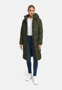 Lungo cappotto imbottito verde oliva con cappuccio, dotato di zip frontale, tasche laterali e polsini a costine, indossato sopra una maglietta bianca e jeans blu.