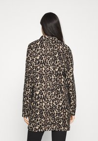 Kobieta z długimi ciemnymi włosami, ubrana w płaszcz w panterkę o średniej długości, stojąca tyłem na jednolitym jasnym tle.