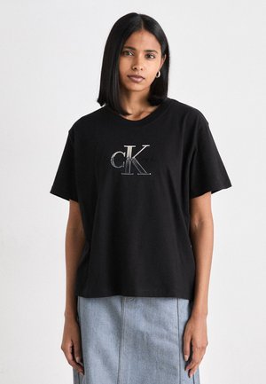 Camiseta de algodón negra con mangas cortas, con un logo en plata "CK" y textura suave. Combinada con una falda de mezclilla.