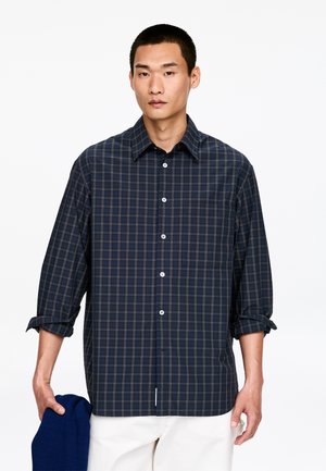 Chemise à carreaux bleu marine à manches longues, avec des boutons sur le devant et des poignets retroussés ; tenue dans une main avec un pull bleu plié.