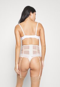 Wit lingerieset met een doorzichtige tailleverkleiner met kantafwerking, verstelbare bandjes en kousenbandbevestigingen. Lichtbeige accenten en texturen.