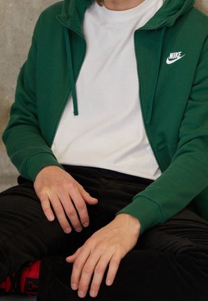 Personne portant un sweat à capuche zippé vert Nike sur un t-shirt blanc, pantalon noir, assise les mains posées sur les genoux.