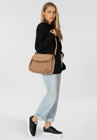 Braune Leder-Schultertasche mit einem Reißverschluss oben, Fronttasche und verstellbarem Schulterriemen, getragen von einer Person in lässiger Kleidung.