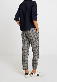 Kvinna som bär en svart topp med trekvartsärmar, svarta och vita rutiga byxor och vita sneakers, står med händerna i fickorna och vänder ryggen åt.