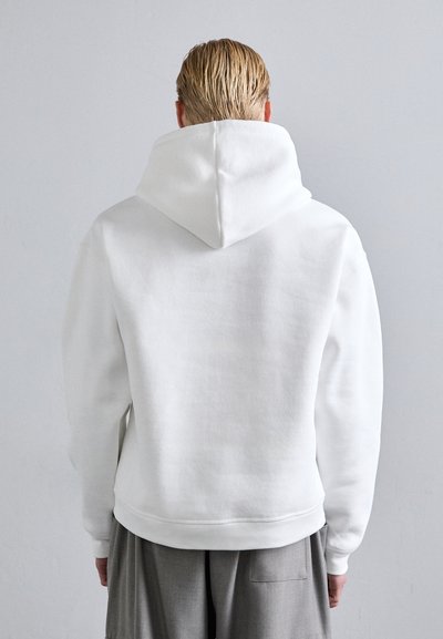 Sudadera blanca con capucha, fabricada con tejido texturizado, puños y dobladillo de canalé. Presenta un corte holgado y una capucha visible en la parte posterior.