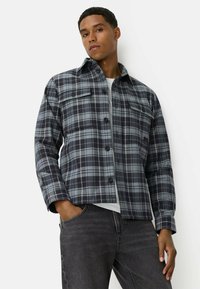 Veste en tartan gris foncé et bleu clair, en tissu, avec deux poches poitrine, fermeture à boutons et un col.
