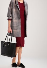 Žena v burgundských šatech a šedém kostkovaném kabátu drží černou tašku, na nohou má černé slip-on boty s chlupatými detaily.