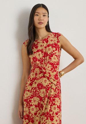 Femeie cu păr lung și întunecat, purtând o rochie roșie fără mâneci cu imprimeu floral bej și detaliu nod răsucit în talie, stând în fața unui perete simplu.