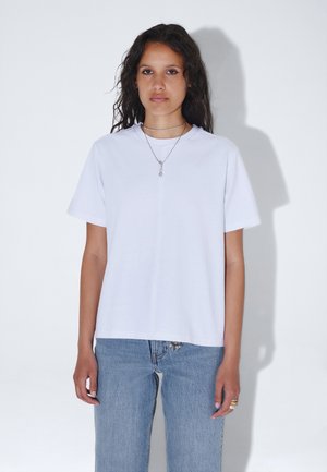 Mujer con pelo largo y oscuro, vestida con una camiseta blanca lisa, vaqueros azules, un collar y anillos, de pie frente a un fondo claro.