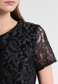 Schwarzes Spitzenkleid mit aufwendigen floralen Mustern, kurzen Ärmeln und einer Schleifenakzent am Auschnitt, mit einem transparenten, strukturierten Finish.
