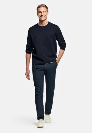 Homme souriant marchant, portant un pull noir, un pantalon foncé et des baskets blanches, une main dans la poche, sur un fond blanc uni.