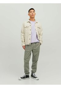 Giacca di jeans beige, t-shirt lavanda e pantaloni jogger verde chiaro. Sneakers alte nere con suola bianca. Outfit casual con design funzionale.
