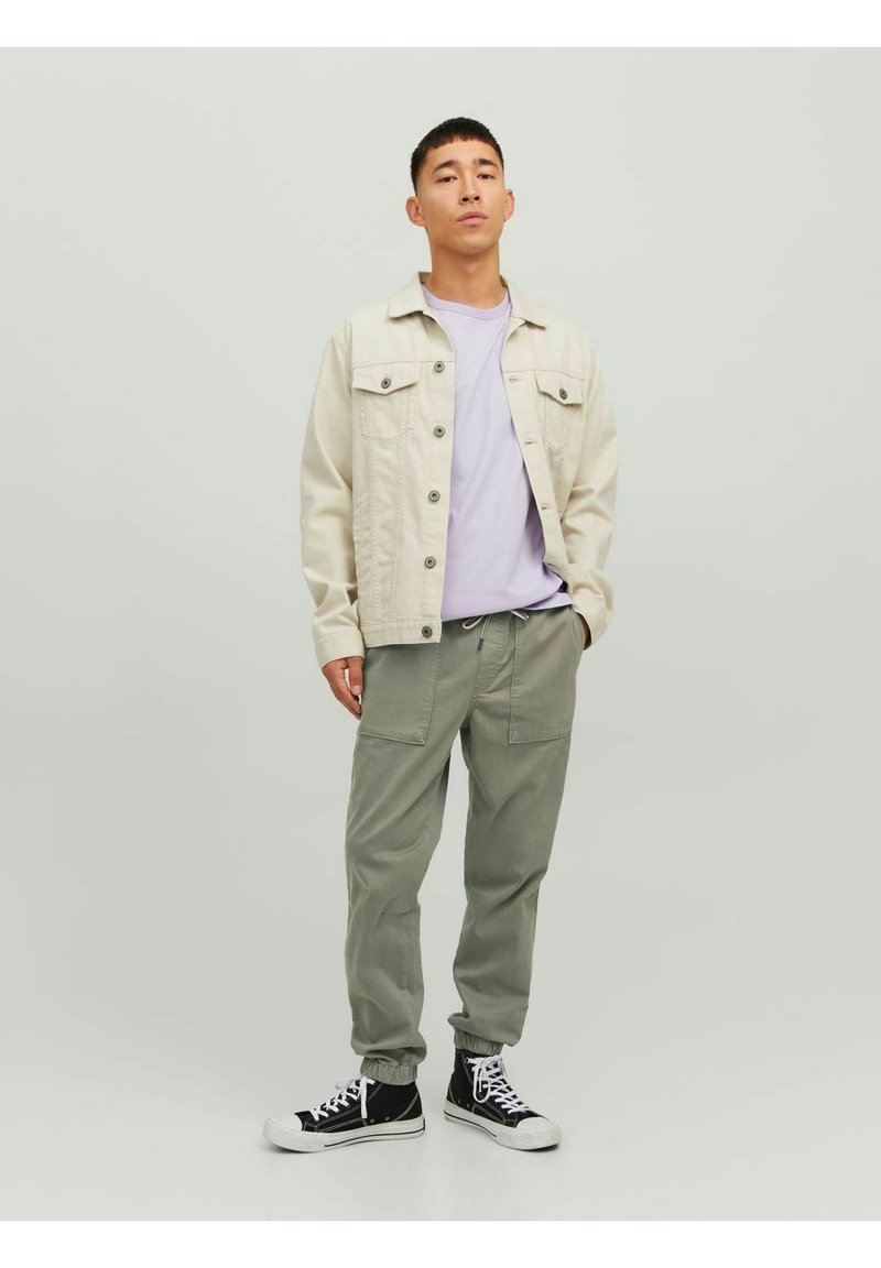 Giacca di jeans beige, t-shirt lavanda e pantaloni jogger verde chiaro. Sneakers alte nere con suola bianca. Outfit casual con design funzionale.