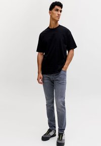 Ung mand stående med den ene hånd i lommen iført en sort t-shirt, grå jeans og grå-sorte sneakers mod en ensfarvet baggrund.