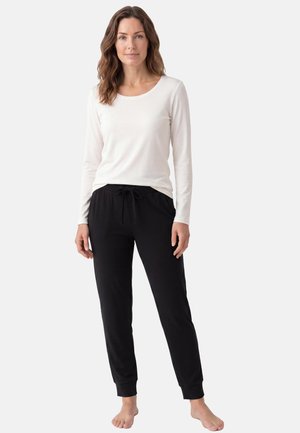 Donna in piedi a piedi nudi che indossa un top bianco a manica lunga e pantaloni neri con coulisse su uno sfondo chiaro uniforme.
