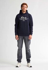 Tmavě modrá mikina s kapucí s bílým nápisem, kapsou typu klokanka, žebrovanými manžetami a lemem. Kombinováno se šedými džínami a bílými teniskami.