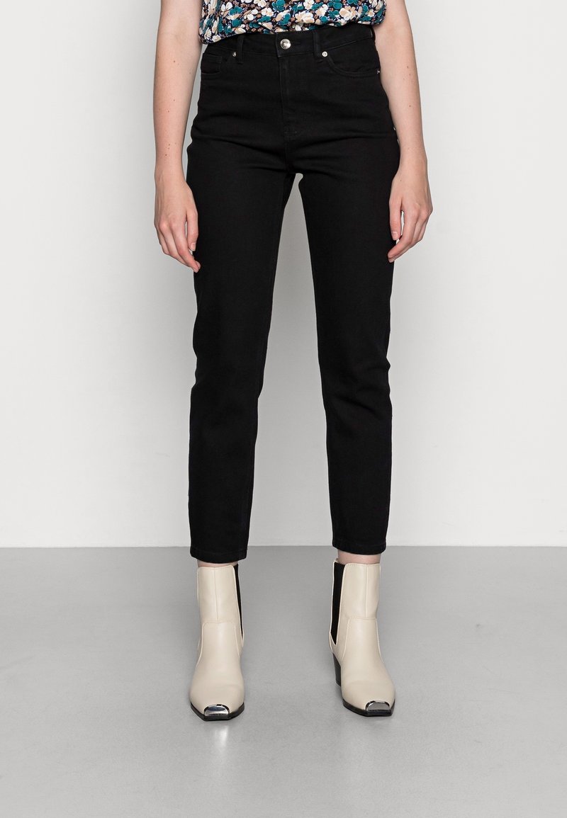 Femme portant un pantalon noir taille haute slim, un top fleuri rentré dans le pantalon et des bottines crème avec embouts métalliques, debout sur un sol gris.