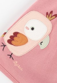 Rosa Stoff mit einem Eulenmotiv und gestickten Details. Die Eule hat ein weißes Gesicht, braune Flügel und grüne Akzente. Textlogo in der Ecke.