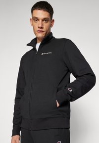 Schwarzer Reißverschluss-Pullover aus weichem Stoff, mit hohem Kragen, Seitentaschen und kleinem gesticktem Logo auf der Brust und dem Ärmel.
