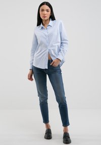 Camicia azzurro chiaro con bottoni in cotone, abbinata a jeans fitted blu scuro e scarpe slip-on nere. Design semplice, maniche lunghe.