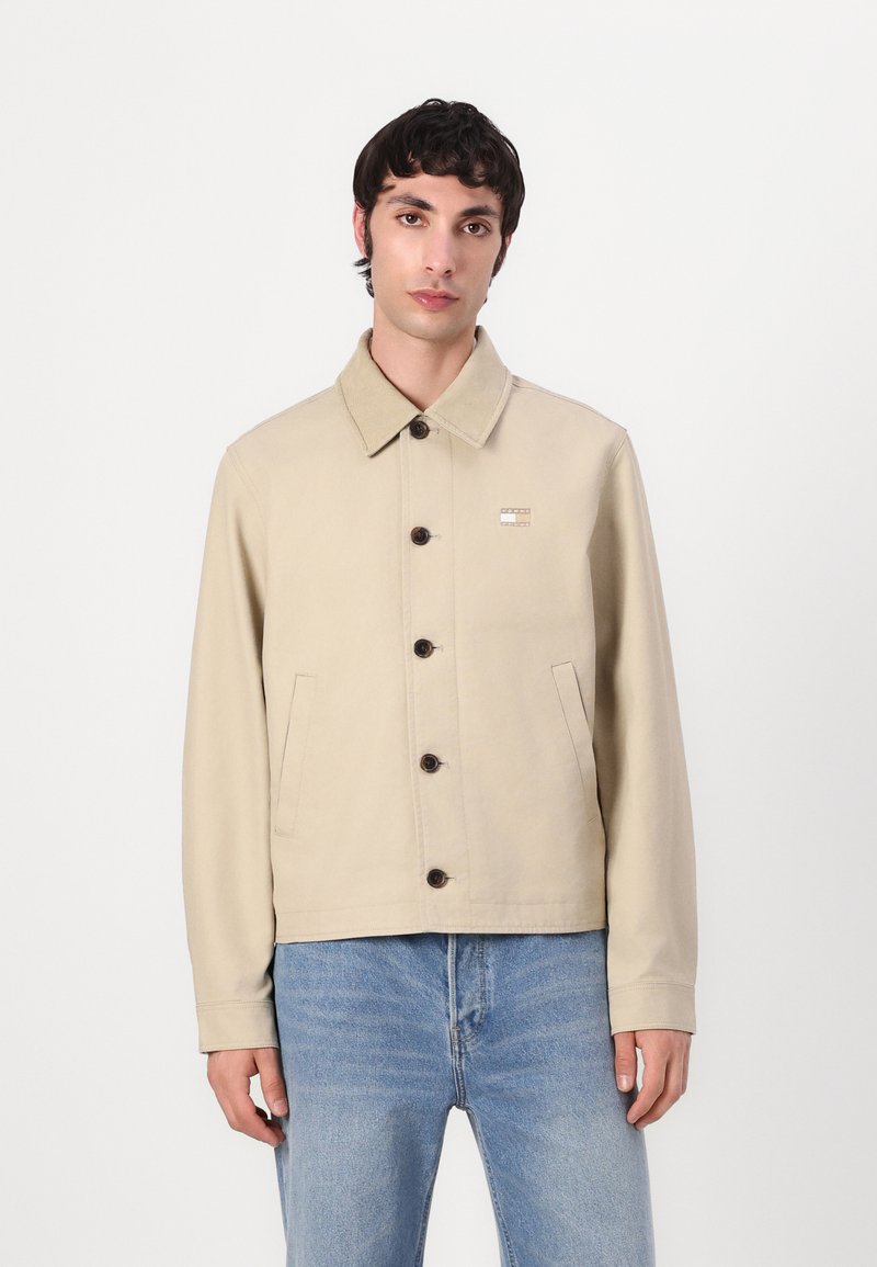 Beige lerretjakke med krage, brune runde knapper, sidelommer og et lite logo-patch på venstre bryst.