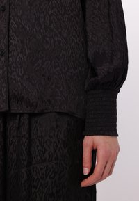 Schwarze Bluse mit strukturierter Oberfläche und dezentem Tiermuster, mit lockerer Passform, Knopfverschluss und gerafften Manschetten.