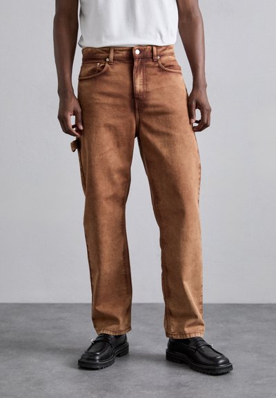 Bruine denimbroek met een relaxte pasvorm, voorzien van vijf zakken, metalen hardware en een soepele textuur, gecombineerd met zwarte schoenen.