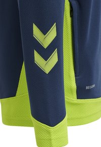 Marineblau und neongrüne Sportjacke aus strukturiertem Material, mit Chevron-Muster an den Ärmeln und einer Reißverschlusstasche.
