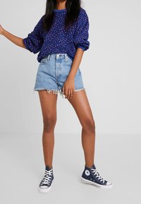Woman wearing blue leopard print sweatshirt, light denim frayed shorts, and navy high-top sneakers standing on a white floor.