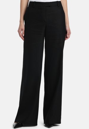 Zwarte broek met wijde pijpen gedragen door een persoon, gecombineerd met puntige zwarte schoenen en een ingestopt wit shirt, tegen een effen lichtgrijze achtergrond.