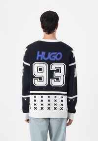 Marine strikkegenser med hvite og blå detaljer, stort "HUGO"-logo, nummer "93," og et geometrisk mønster i kanten.