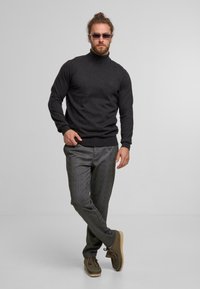 Mann mit Sonnenbrille trägt einen dunklen Rollkragenpullover, graue karierte Hose und braune Wildlederschuhe, steht mit überkreuzten Beinen vor einem schlichten Hintergrund.