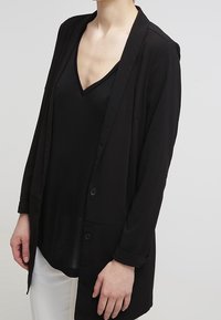 Persona vestida con una camisa negra de cuello en V debajo de un blazer negro con botones, combinada con pantalones blancos, de pie frente a un fondo gris claro.