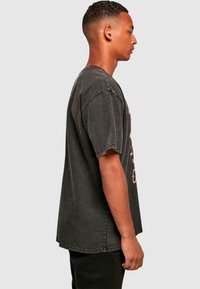 Tricou negru supradimensionat cu guler rotund, mâneci scurte și margini uzate. Prezintă grafica estompată în culori deschise pe față.