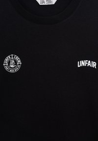 Melnas kokvilnas t-krekls ar baltu apļu logotipu kreisajā krūtī un vārdu "UNFAIR" baltā krāsā labajā pusē.