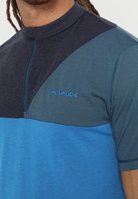 Kratkým rukávem cyklistické tričko s čtvrtinovým zipem, spojující tmavě námořnickou a gradientní modré panely, s jemně strukturovaným povrchem a logem.