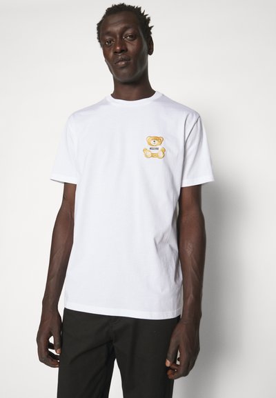 Camiseta de algodón blanca con un pequeño diseño de osito de peluche bordado en marrón en el pecho. Estilo de mangas cortas y cuello redondo, con una textura suave.